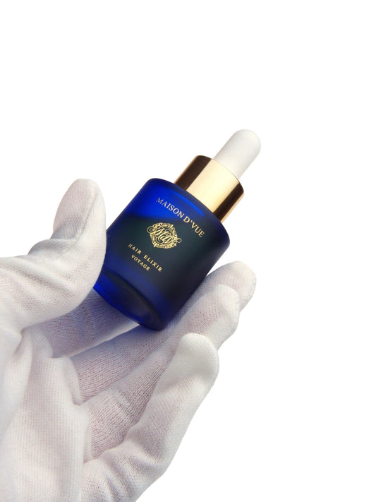 A white-gloved hand holds the 10ml VOYAGE bottle of MAISON D'VUE Hair Elixir in frosted cobalt glass with a gold collar, against a pure white background.